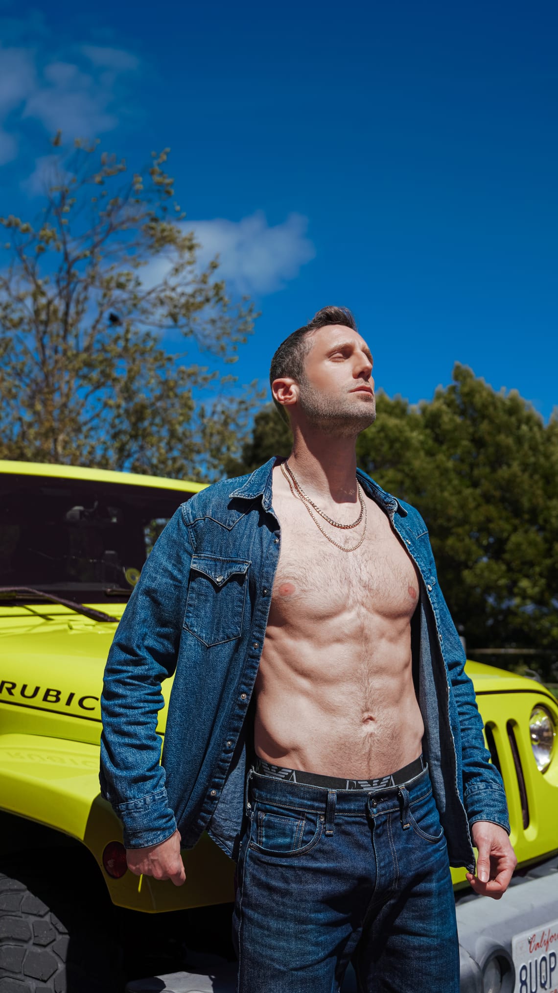 Model standing in front of a green Jeep wearing an open denim western-style shirt